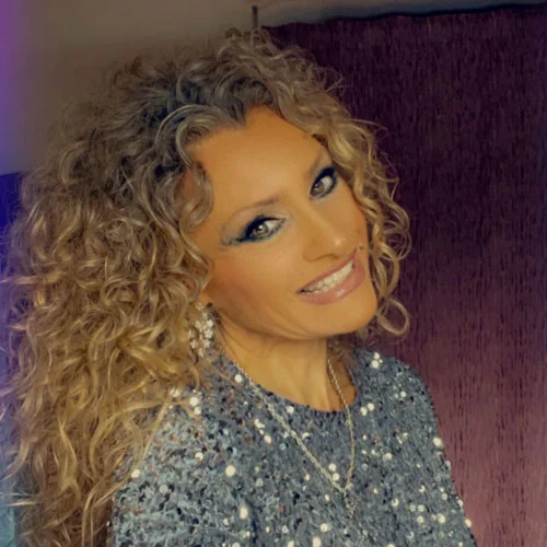 This is a portrait of a woman with a radiant smile and curly blonde hair, wearing a sparkling sequined top that adds a dash of glamour. Her joyful expression exudes confidence and a positive energy, befitting Kris Saint Ange, a coaching expert dedicated to guiding individuals toward personal and professional fulfillment.